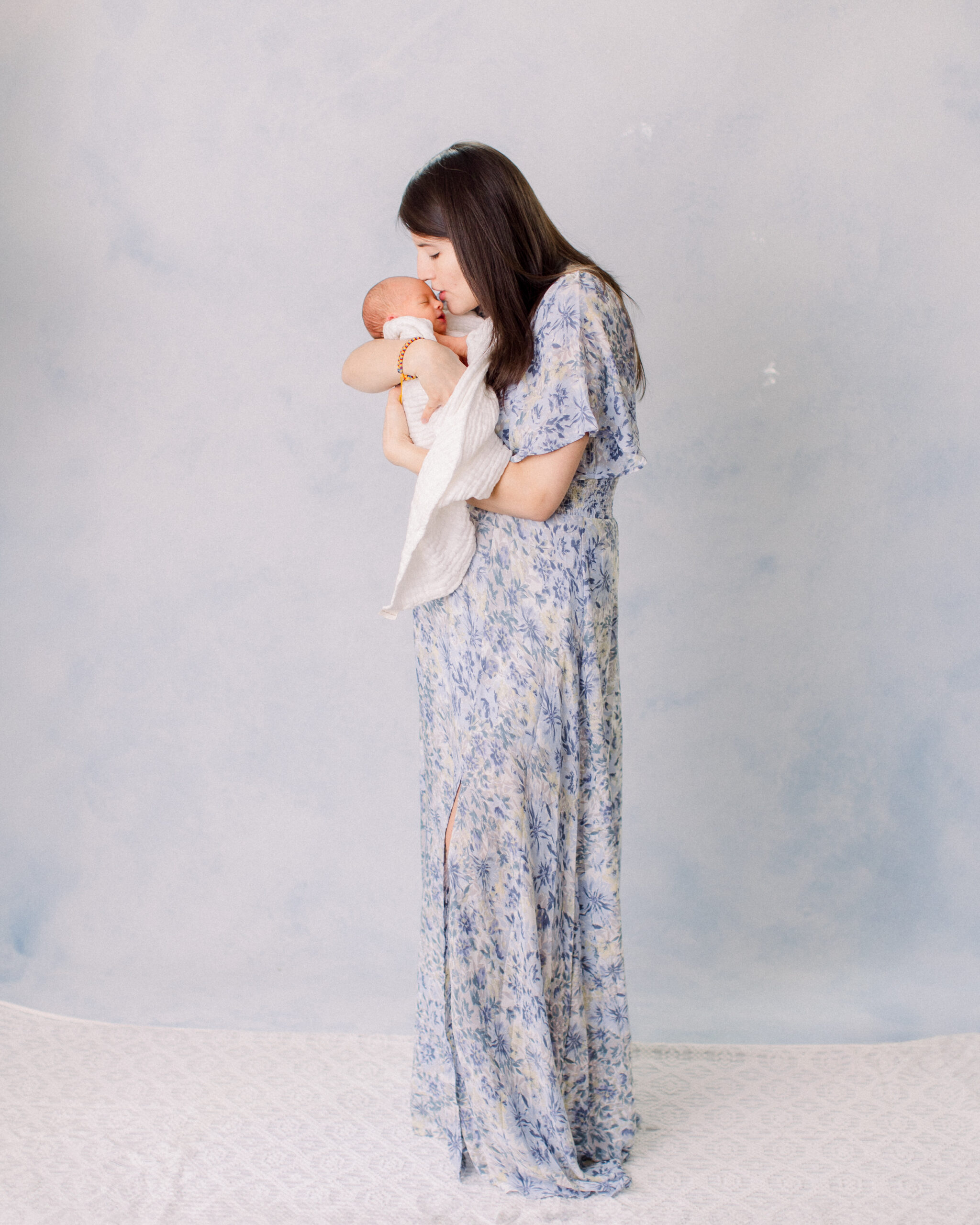 mother in blue dress holding newborn wrapped in white muslin wrap, standing in front of a blue hand painted backdrop