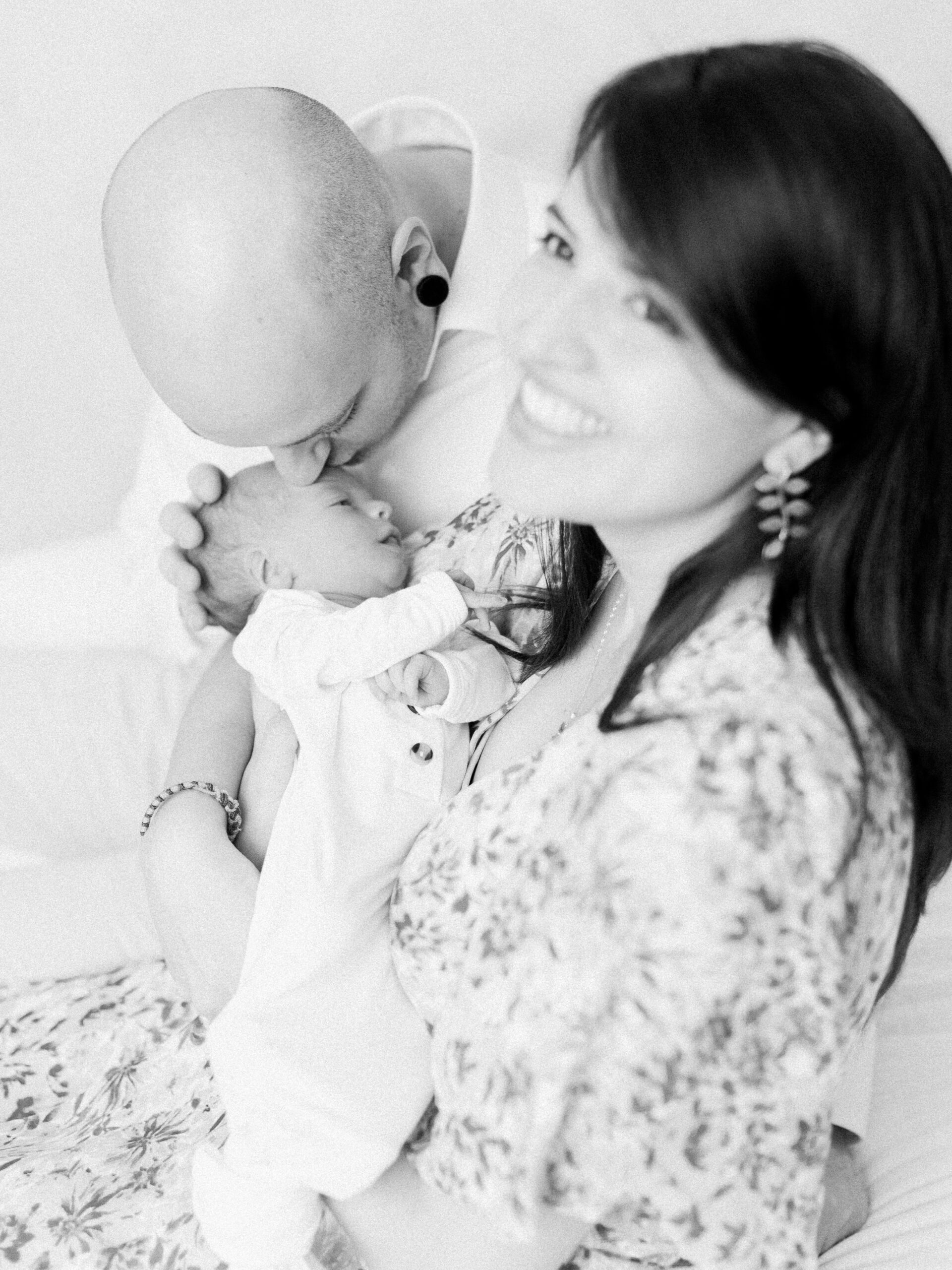 Mother and father holding newborn baby, mother smiling up and father leaning down to kiss the baby's head.