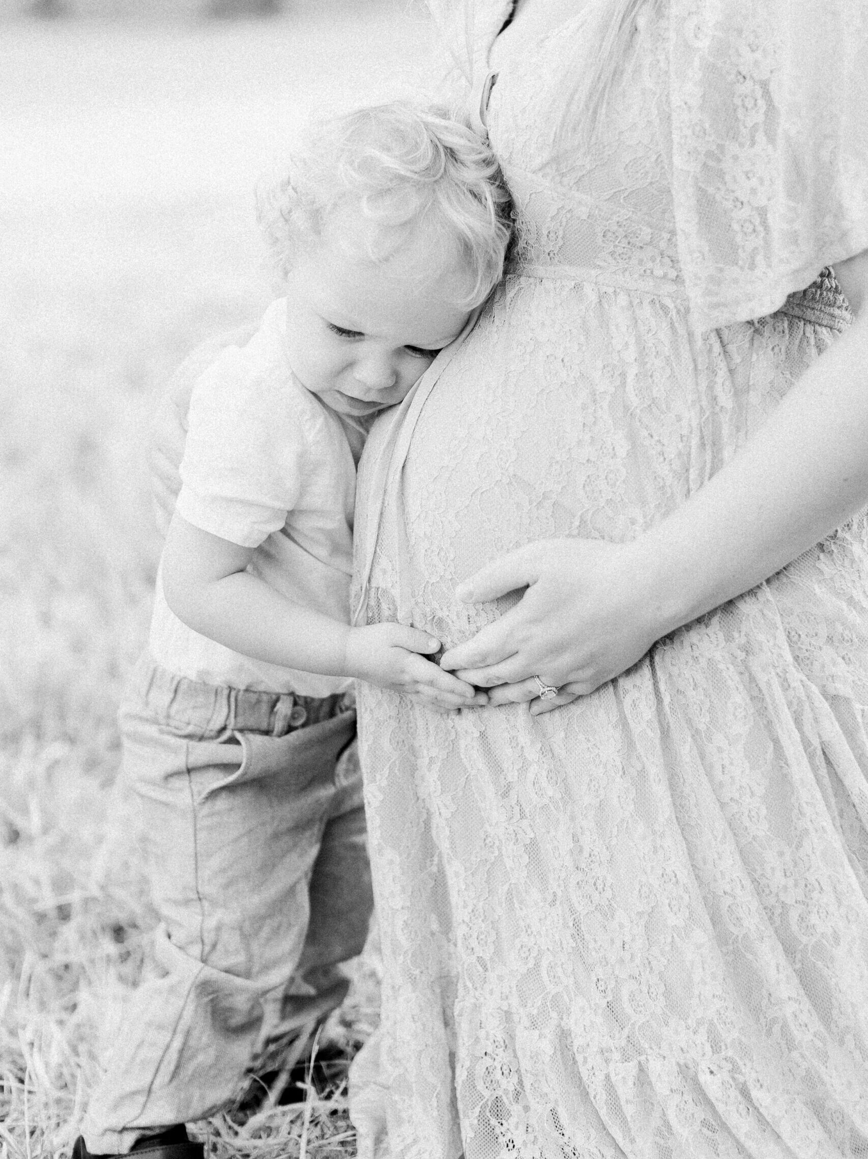 Toddler boy embracing mom's baby bump in an open field.