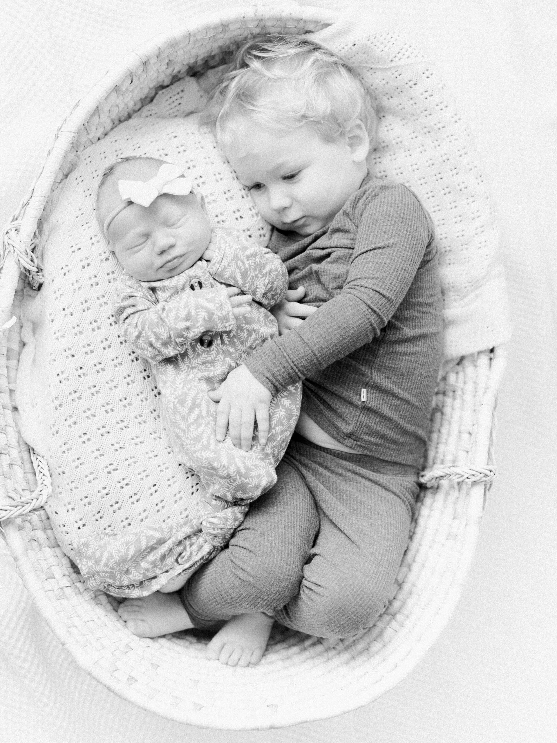 Two year old brother laying down next to newborn baby sister in a wicker basket, giving soft snuggles.