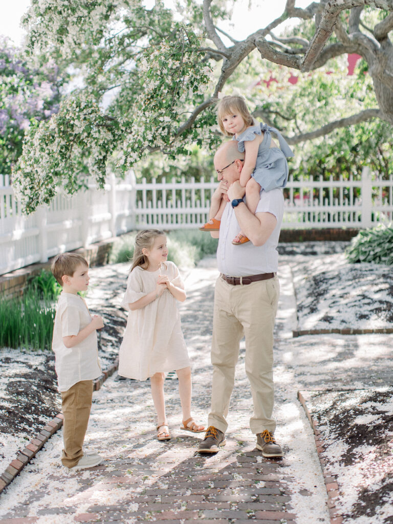 Family in a floral filled, ethereal garden with toddler on dad's shoulders and older siblings gathered around.