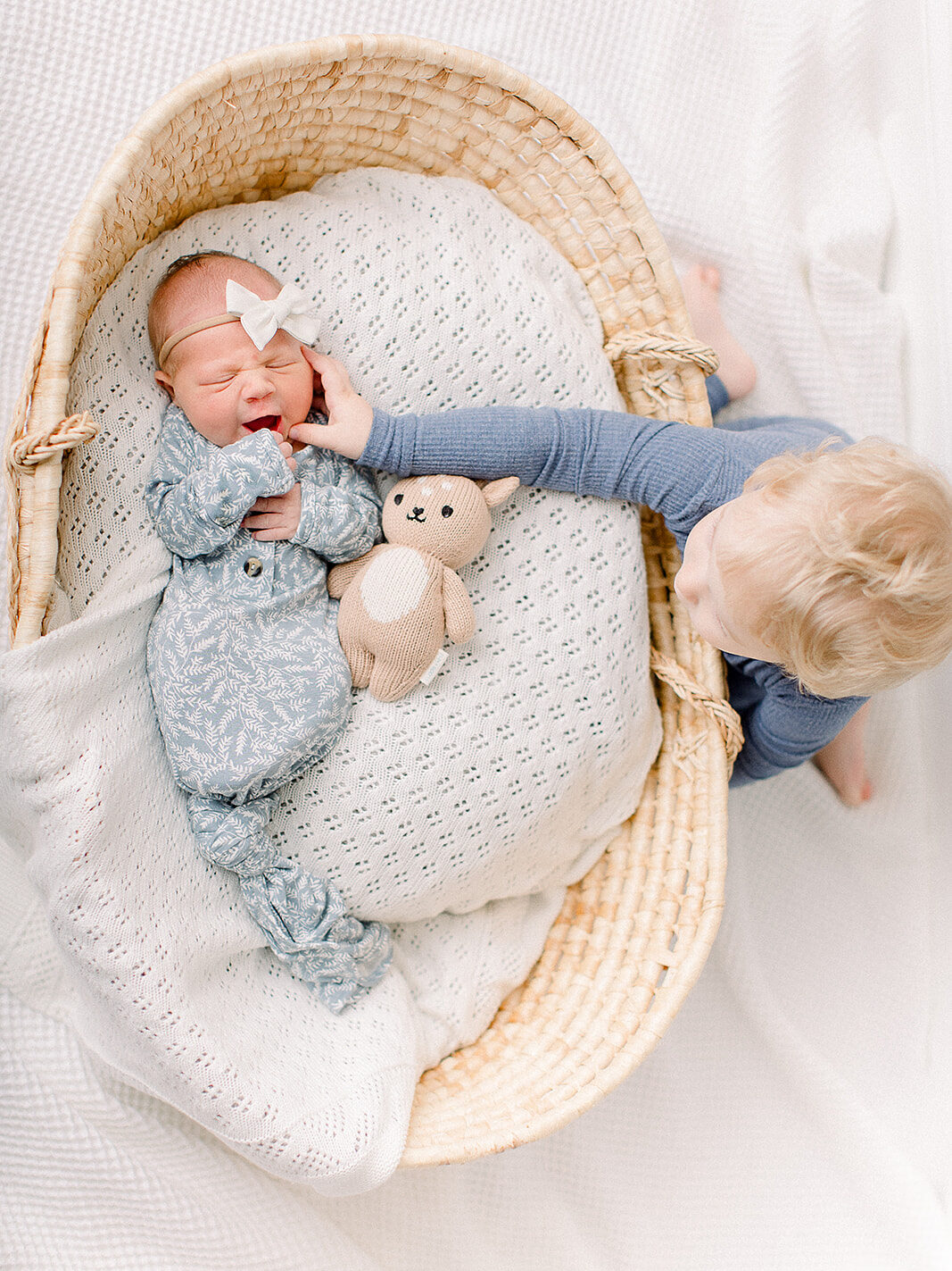 Older sibling gently touching newborn sister’s face as she rests in a woven basket during a cozy at-home newborn session. By Boston & Northshore Newborn Photographer Fieldstone Studio