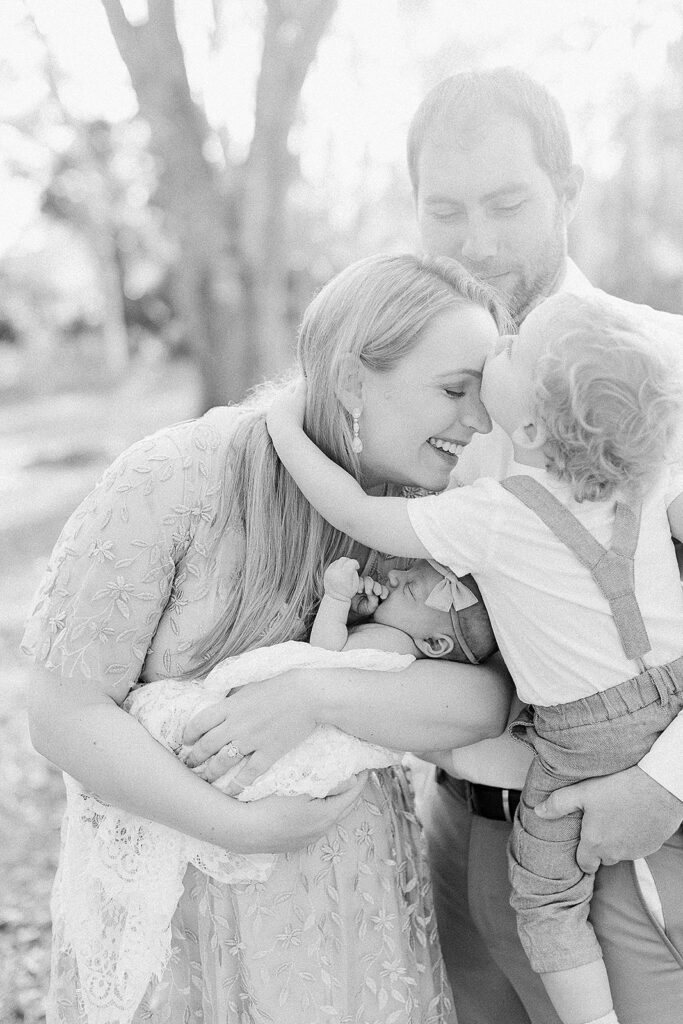 Mother smiles as her toddler kisses her forehead while she cradles newborn baby in arms during a heartfelt family newborn session. By Boston & Northshore Newborn Photographer Fieldstone Studio