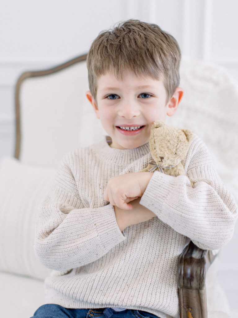 Little boy sitting in a white French armchair, smiling and snuggling his teddy bear. Timeless child portraits in Massachusetts by Fieldstone Studio.